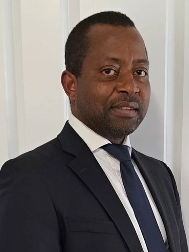 A man in a dark suit and tie posing indoors against a light-colored wall.