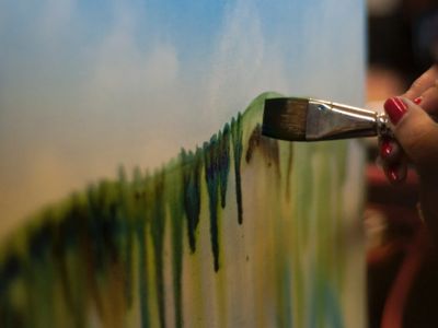 A hand painting a landscape with a brush on canvas.