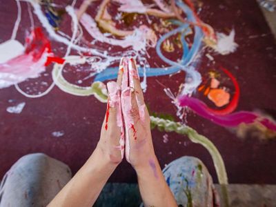 Hands covered in colorful paint held together in a prayer-like pose over a paint-splattered surface.