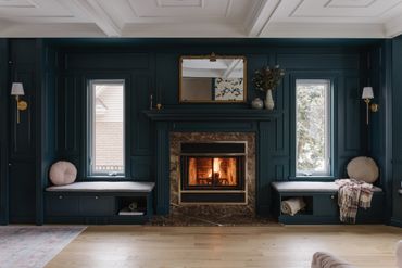 Cozy living room with a lit fireplace, dark walls, and symmetrical seating by the windows.