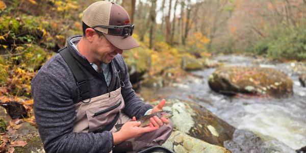 Man holding a small fish by a river.