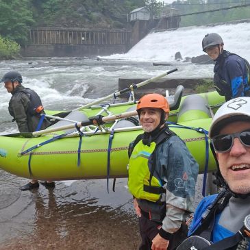Four men with helmets and life jackets carrying a raft by a river.