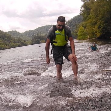 Man wading through a river with backpack and sunglasses.