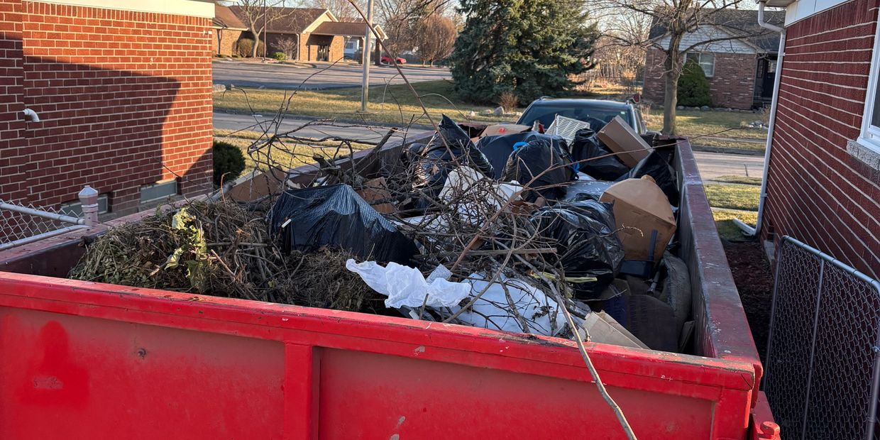 Red dumpster filled with yard waste, branches, and black garbage bags.