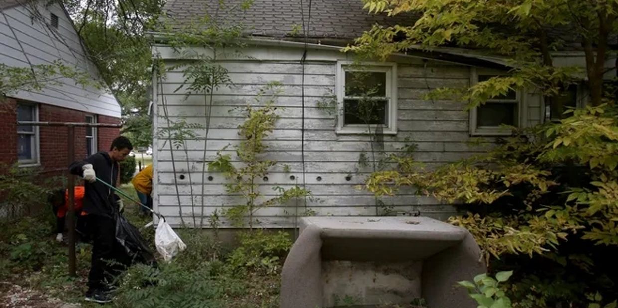 Two people cleaning up an overgrown yard beside a weathered house.