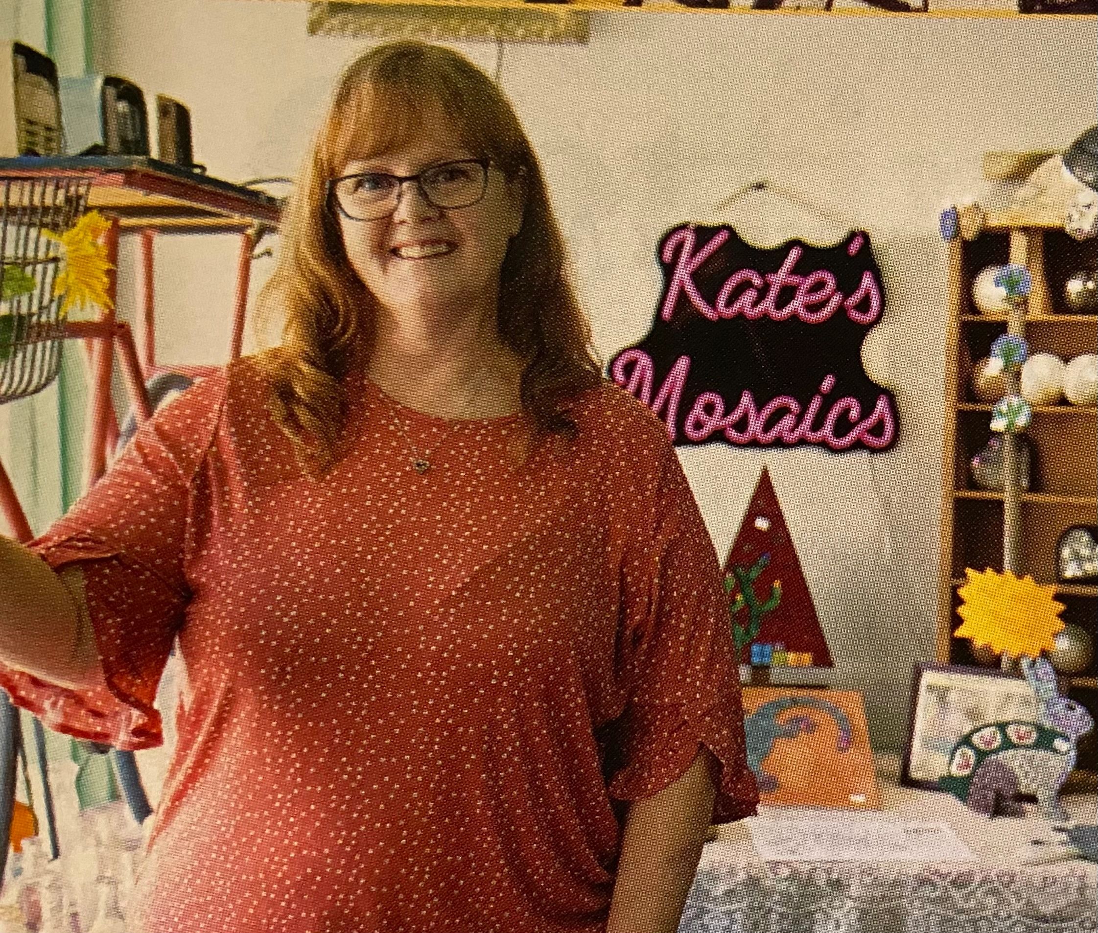 Kate standing in front of mosaics pieces and Kate's Mosaics sign.