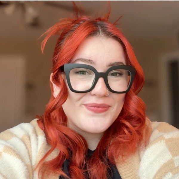 Young woman with vibrant red hair and large black glasses smiling. Esthetician North Branch