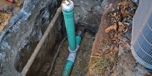Trench with green plumbing pipes installed underground beside an air conditioning unit.