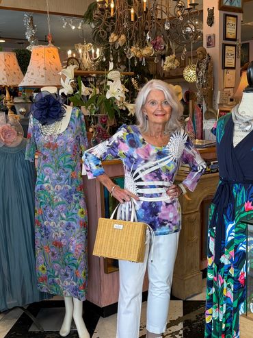 Stylish older woman posing in a boutique with colorful dresses and accessories.