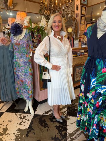 Woman in white pleated dress posing in a boutique with colorful dresses.