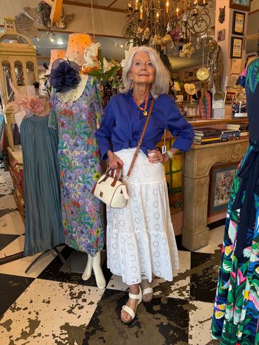 Elderly woman in a white dress and blue jacket posing indoors surrounded by colorful dresses and vintage decor.