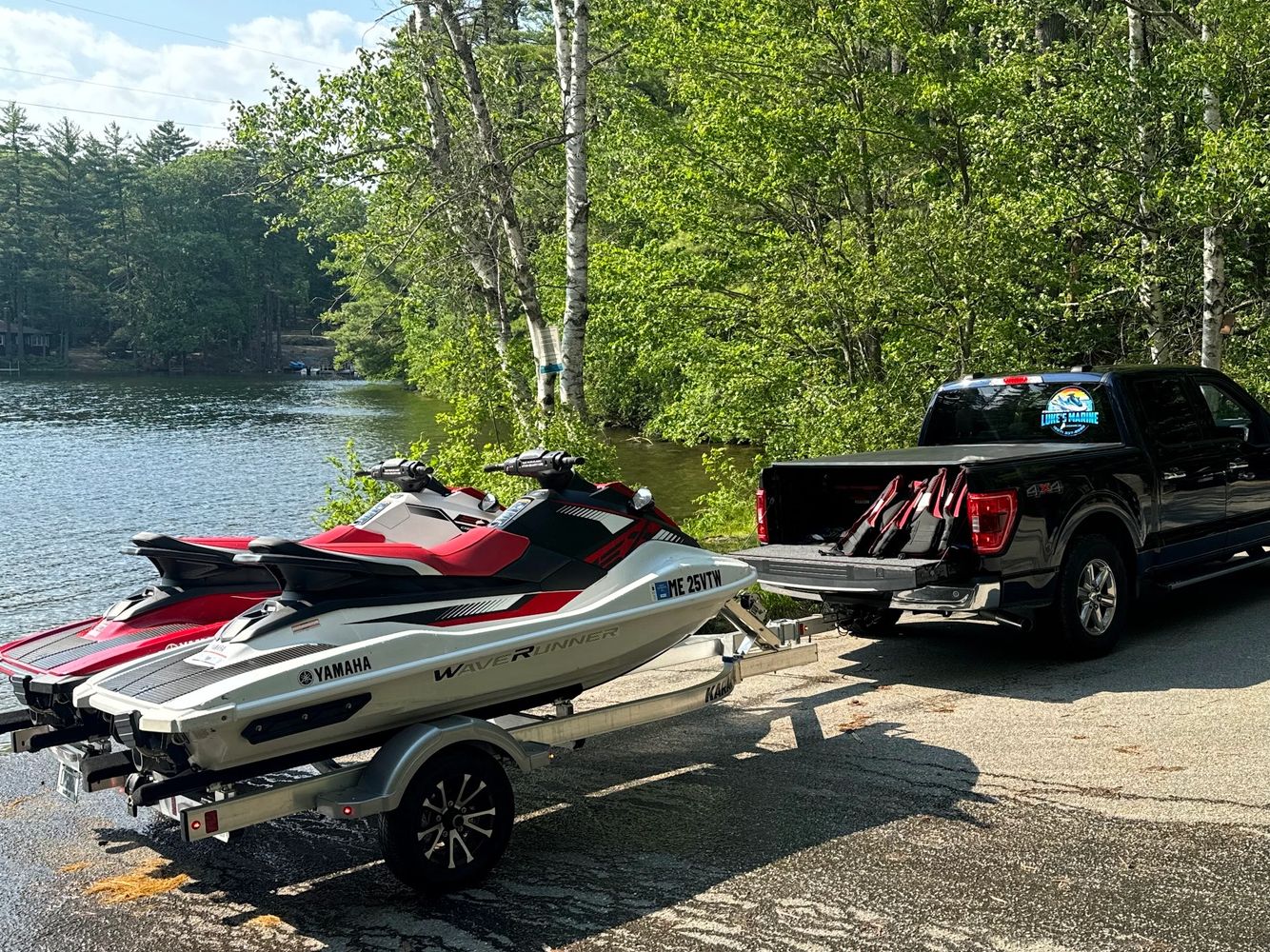 Two Yamaha WaveRunner jet skis on a trailer hitched to a black pickup truck by a lakeside.