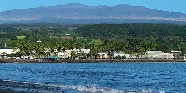 Coastal town with palm trees, ocean waves, and distant mountains under a clear blue sky.