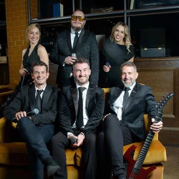 A stylish band dressed in black posing with microphones and a guitar on a yellow couch.