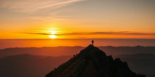 A person stands atop a mountain peak at sunset with vibrant orange skies.