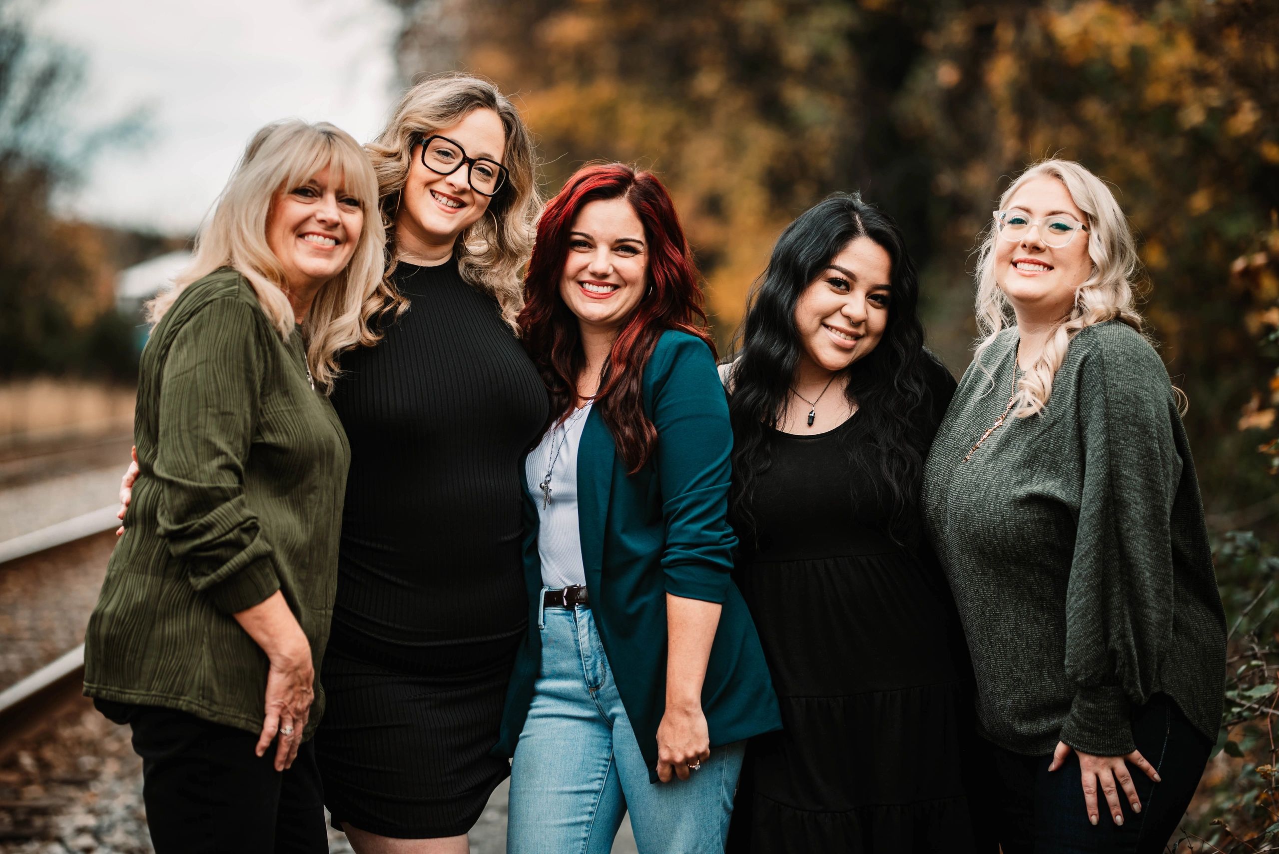 Five women smiling together outdoors near train tracks in autumn.