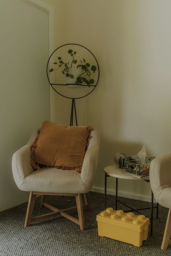 Cozy corner with a beige chair, rust pillow, green plant, and yellow toy box.