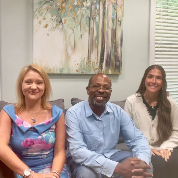 Three people smiling on a couch with a forest painting behind them.