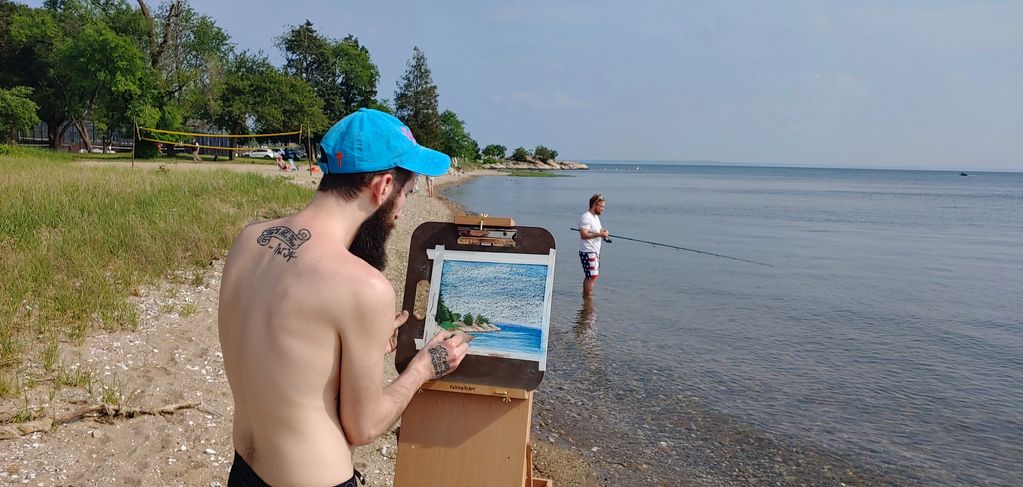 A man paints a beach scene while another fishes in the water.