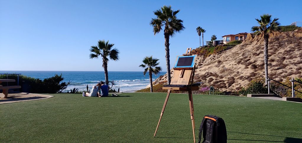 An easel and backpack on grass near a beach with a couple sitting in the distance.