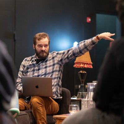 Man in plaid shirt with laptop gesturing during a discussion.
