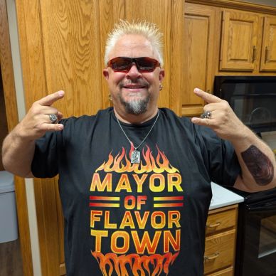 Man in sunglasses wearing a 'Mayor of Flavortown' shirt making rock hand signs.