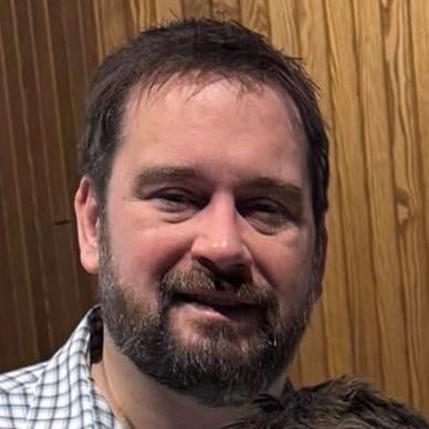 Man with beard smiling indoors against wooden wall.