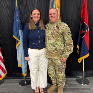 A woman and a soldier in uniform posing in front of flags indoors.