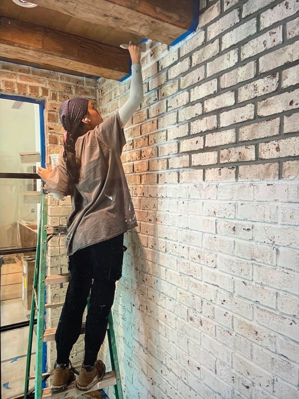 Person on ladder painting a rustic brick wall near wooden beams.