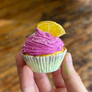 Hand holding a cupcake with purple frosting and a lemon slice on top.