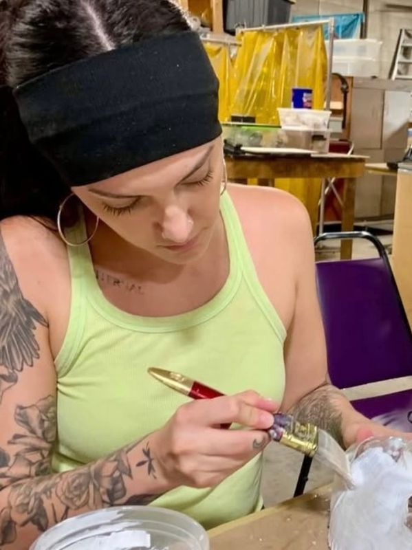 Woman with tattoos painting on a glass jar in a workshop.