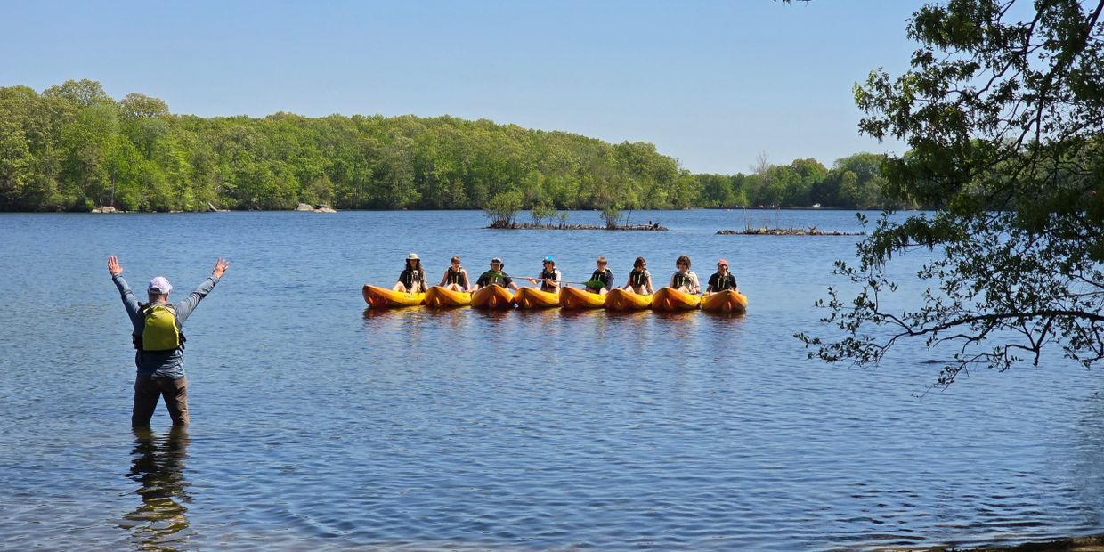 Group kayaking on a calm lake with instructor signaling from the shore.