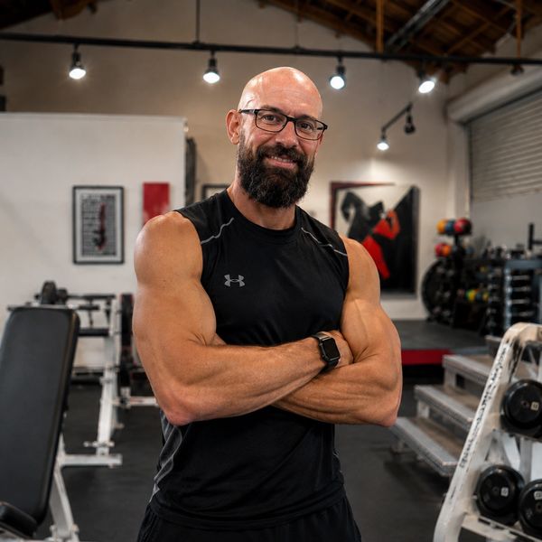 Smiling muscular man with glasses in a gym, arms crossed confidently.