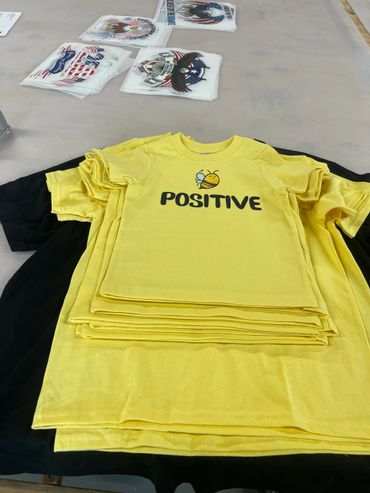 Stack of yellow T-shirts with a bee and the word 'POSITIVE' printed.