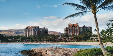 Resort buildings by a beach with palm trees and a person walking on rocks.