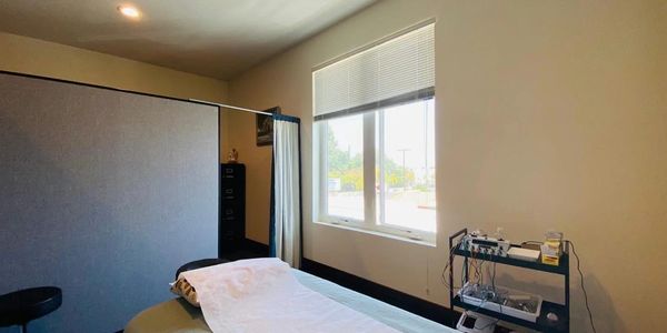 A treatment room with a massage table, stool, and medical equipment near a window.
