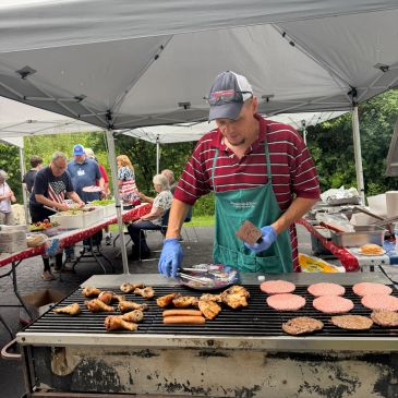 Man grilling meat at outdoor picnic under a canopy.
