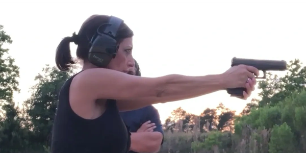 Woman aiming a handgun at an outdoor shooting range with ear protection.