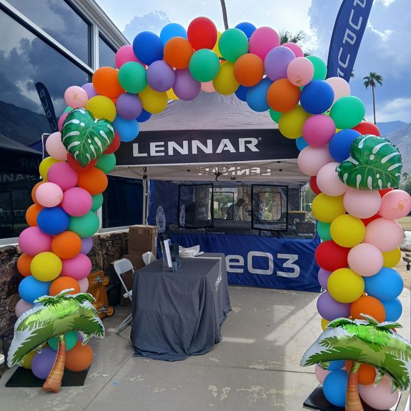 Colorful balloon arch with tropical leaf accents at an outdoor event booth.
