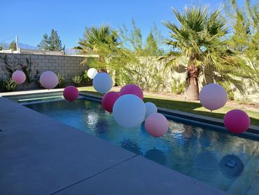 Colorful balloons float above a backyard swimming pool on a sunny day.