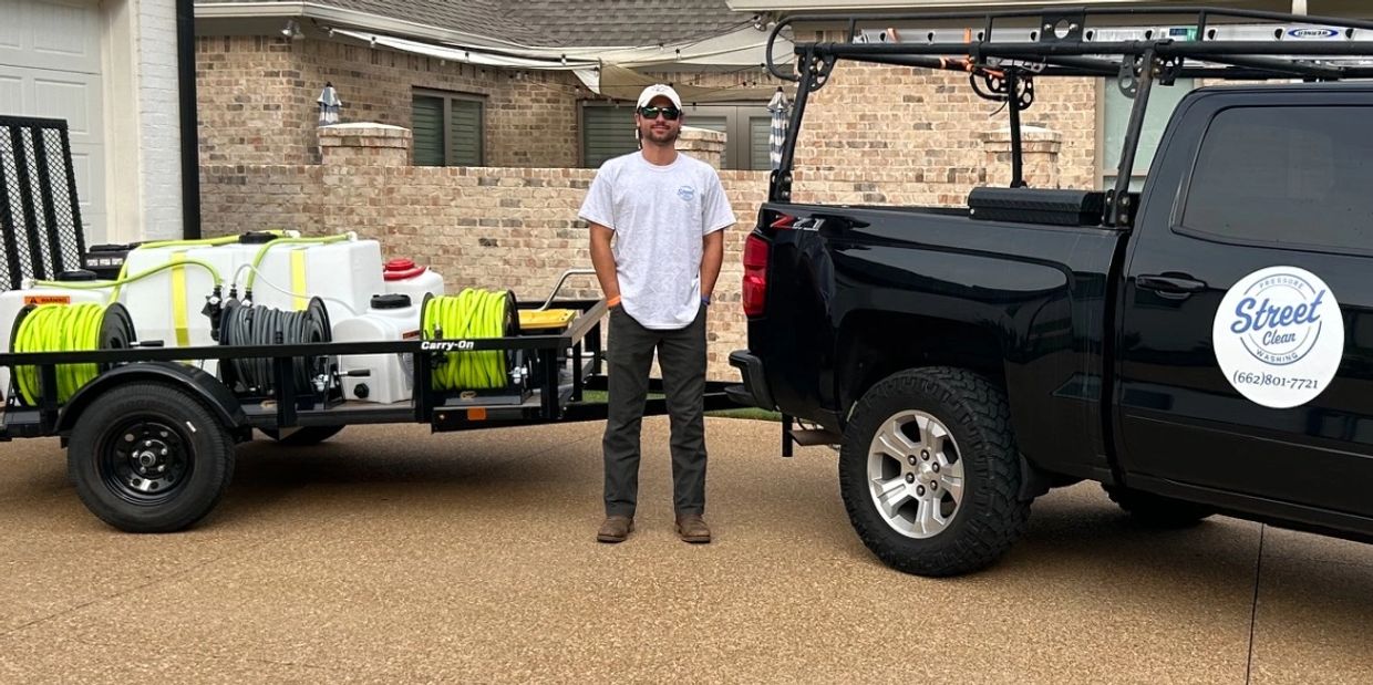 Man standing between a black truck and a trailer loaded with equipment on a driveway.
