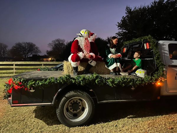Santa and an elf talk with a child on a decorated truck bed at dusk.