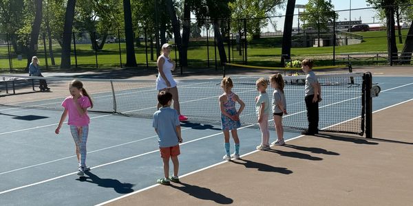 Children lined up on a tennis court with an instructor on a sunny day.