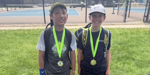 Two boys with medals and tennis gear standing on grass near tennis courts.