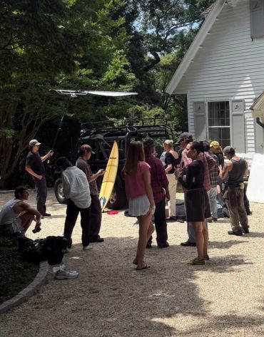 Group of people gathered outside a white house with a surfboard and equipment.