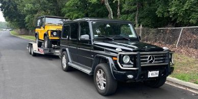 Black Mercedes towing yellow Land Rover on trailer.