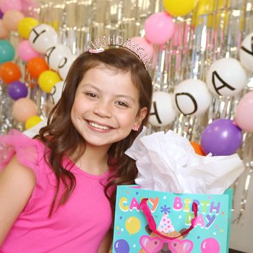 Smiling girl in a pink dress celebrating her birthday with balloons and gifts.