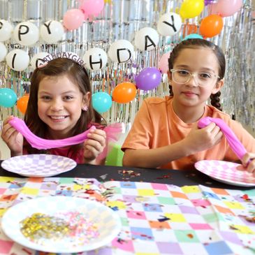 Two girls playing with pink slime at a birthday party with colorful balloons and decorations.