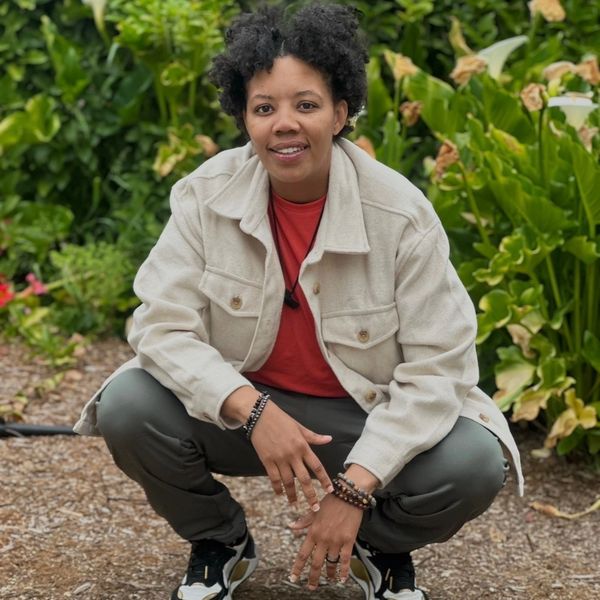 Person squatting outdoors, wearing a beige jacket and sneakers, smiling at the camera.