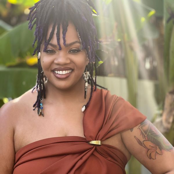 Smiling woman with dreadlocks and a brown wrap dress in natural sunlight.